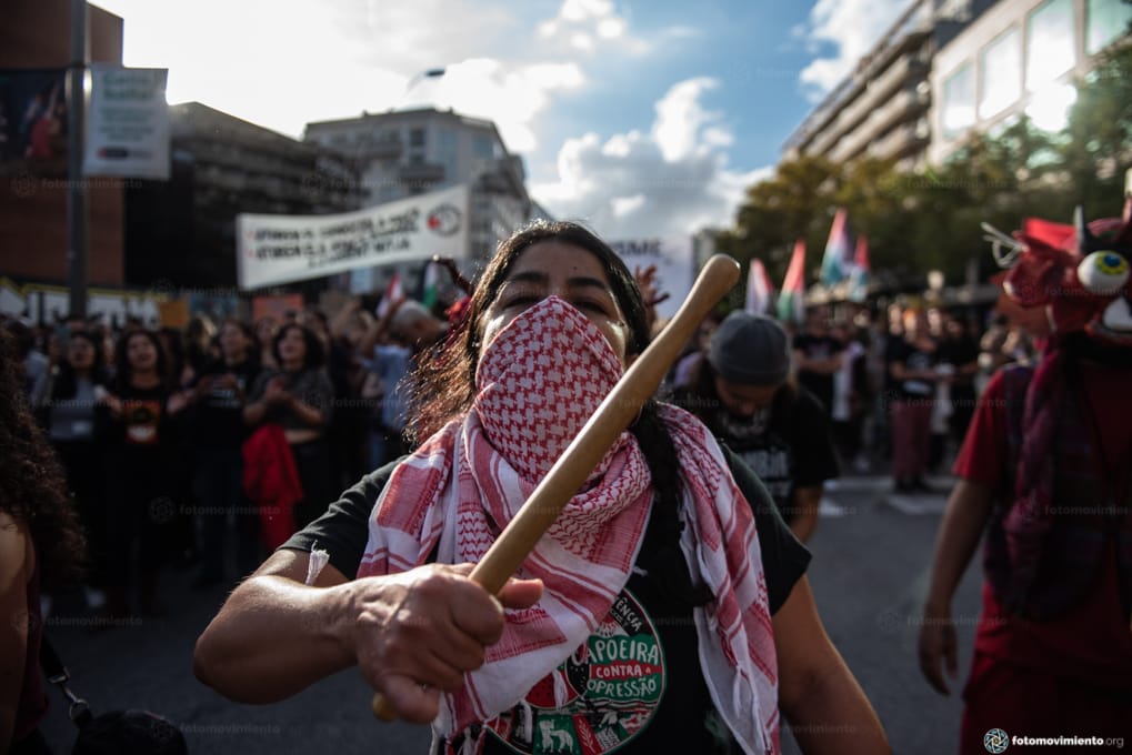 Una manifestante en primer plano con un pañuelo palestino toca un tambor. De fondo le sigue la manifestación. Crédito fotomovimiento.
